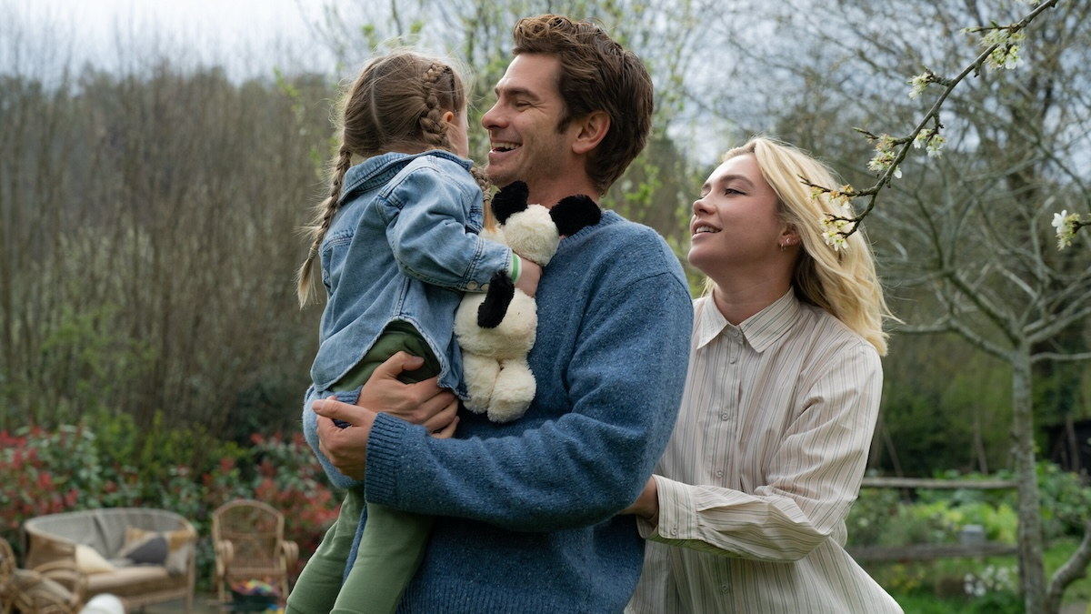 Andrew Garfield i Florence Pugh w scenie z filmu „Sztuka pięknego życia”, trzymają dziecko i uśmiechają się w plenerze.