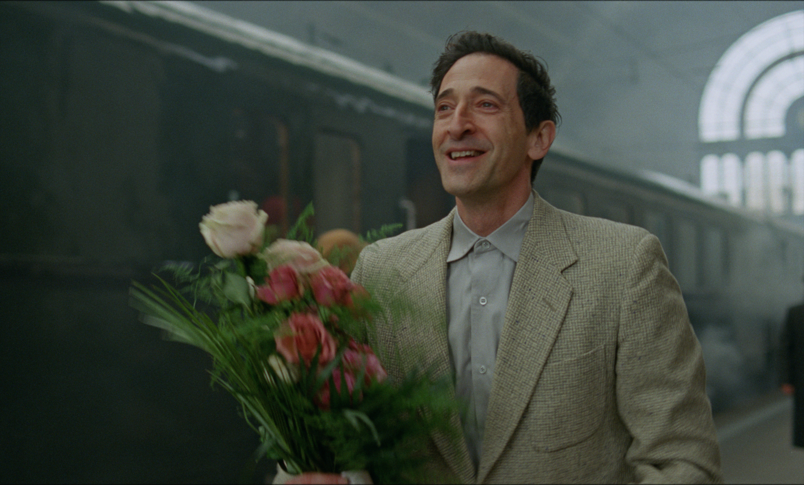 Adrien Brody w filmie „The Brutalist”, z uśmiechem trzyma bukiet róż na dworcu kolejowym.