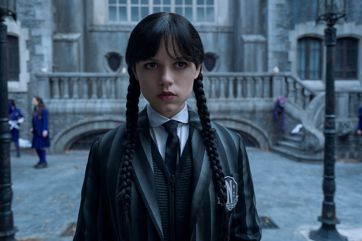 Jenna Ortega jako Wednesday Addams w drugim sezonie serialu Netflix „Wednesday”, stoi na dziedzińcu w mrocznym otoczeniu.