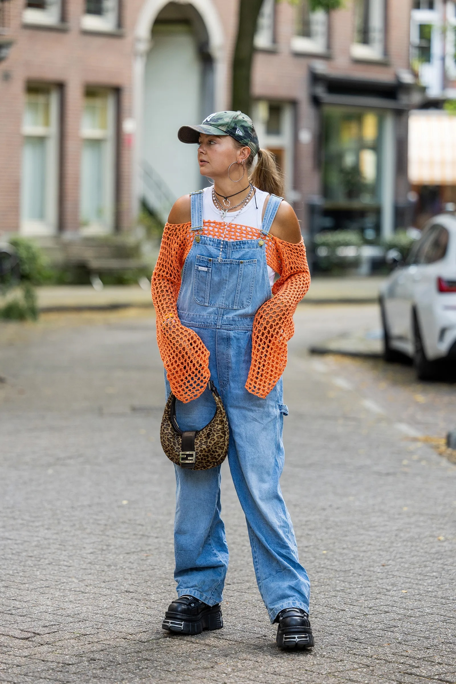 Kobieta w pomarańczowej bluzce szydełkowej boho, jeansowych ogrodniczkach damskich i czarnych crocsach na platformie.