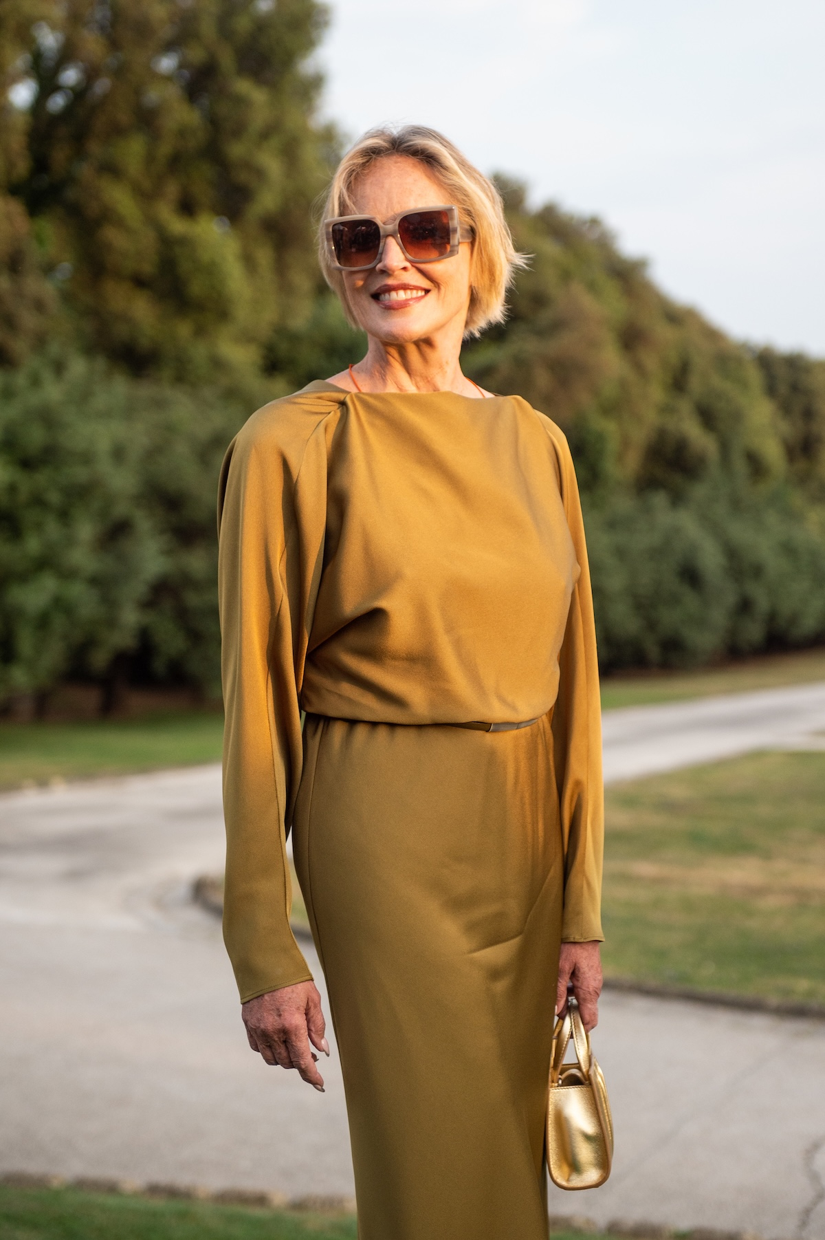 Sharon Stone w sukience na długi rękaw, trzyma złotą torebkę, uśmiecha się w plenerze.