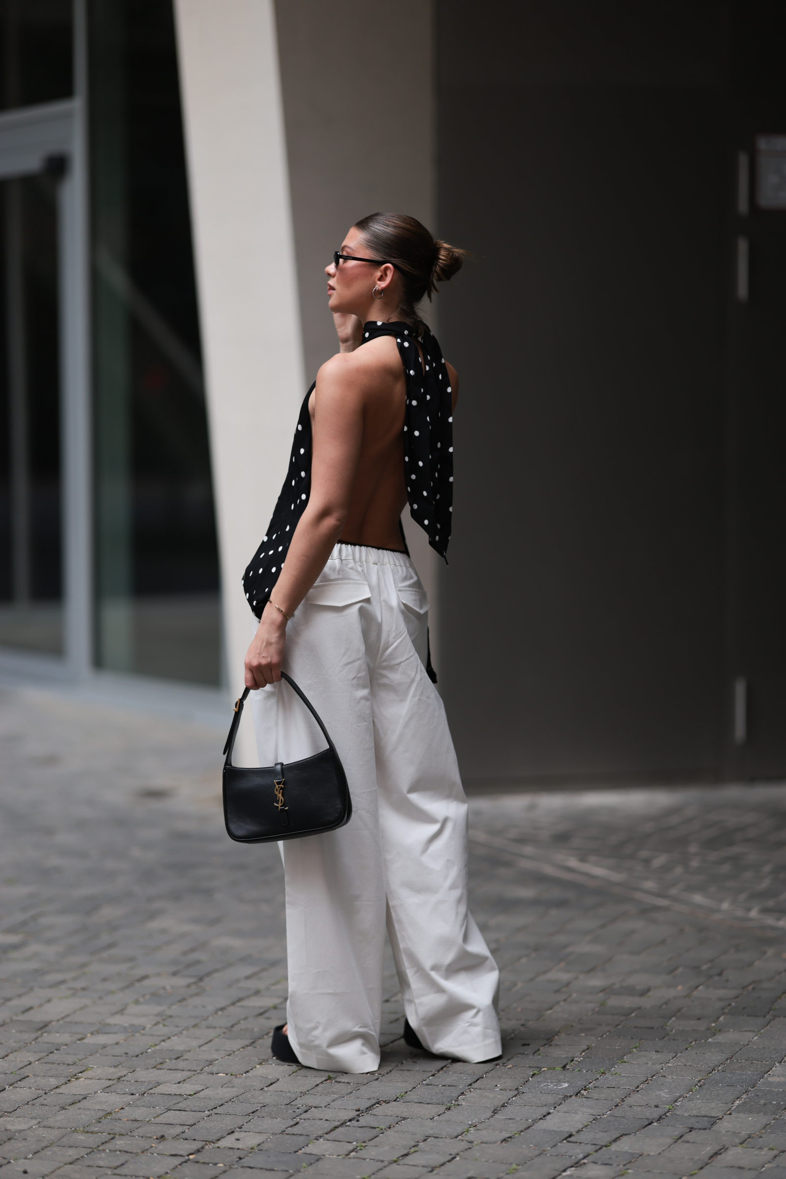 Stylowy outfit z topem eksponującym plecy i białymi spodniami, dodatkiem czarnej torebki.