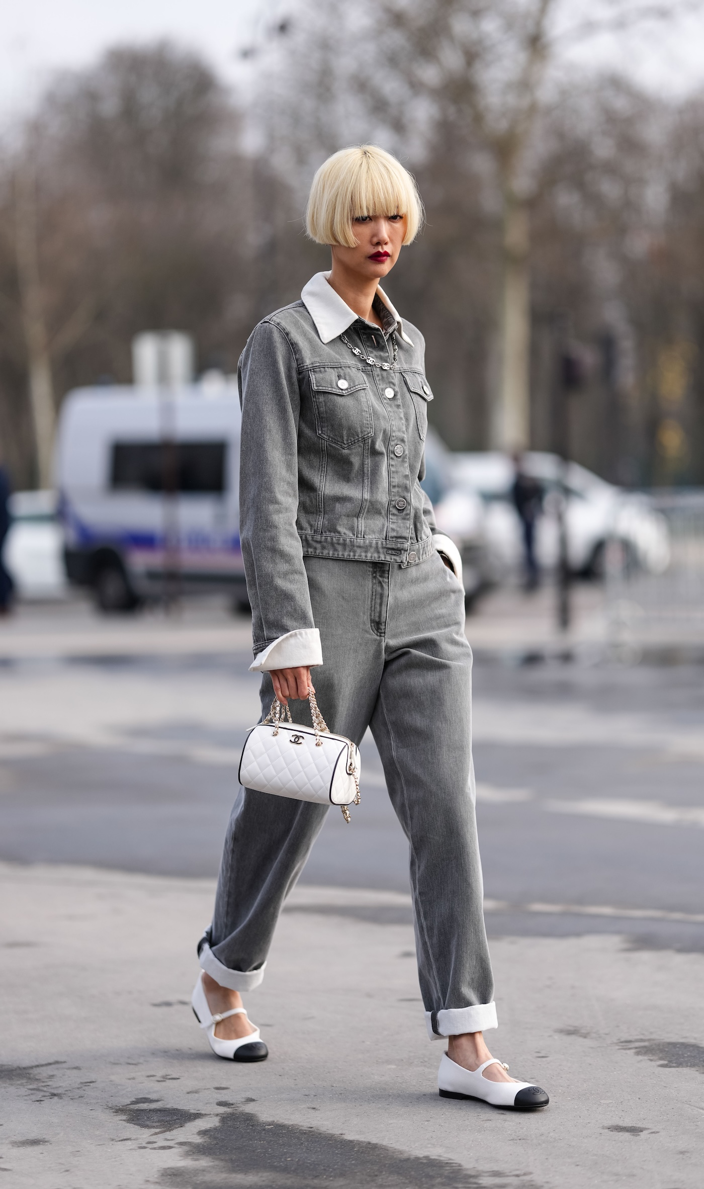 Kobieta sfotografowana podczas Paris Fashion Week, ma na sobie czarno-białe balerinki Mary-Jane oraz jeansowy total look w jasnoszarym kolorze, w ręku trzyma małą białą torebkę Chanel.